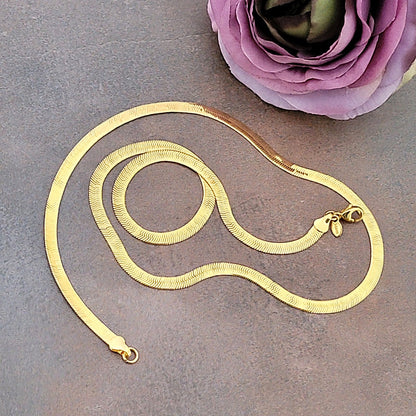 Gold flat chain necklace next to a purple flower.