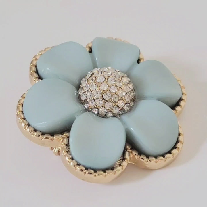 Video of a light blue plastic flower pin, with domed rhinestone center, and gold tone setting..