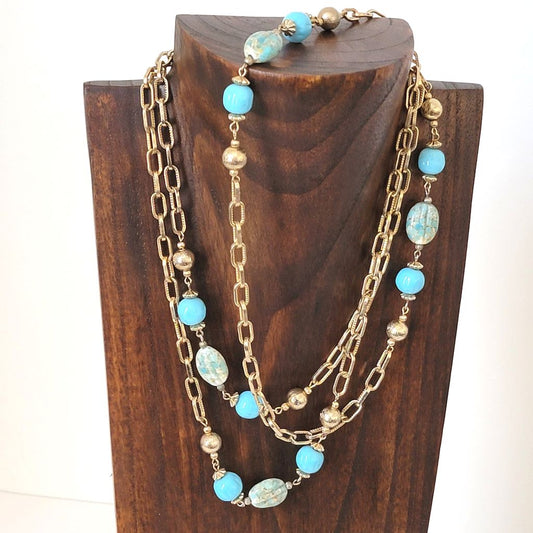 Vintage long gold tone chain necklace with robin's egg blue glass bead accents, shown on a wooden display stand.